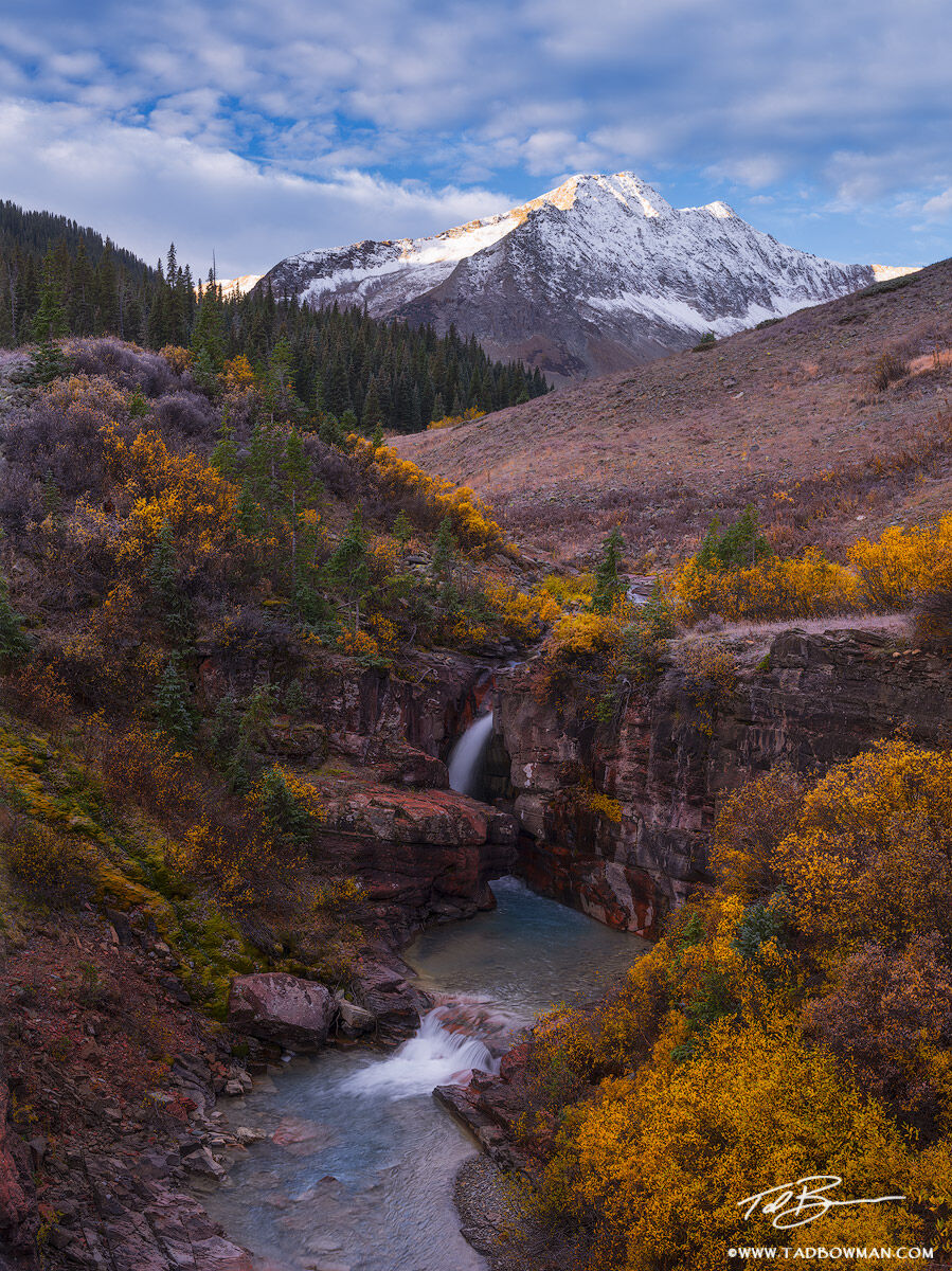 This Colorado mountain photo depicts early morning with a snow capped mountain, waterfall, and fall foliage situated in the San...